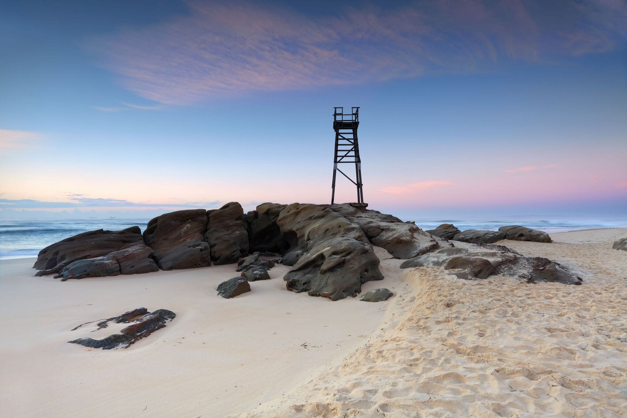 Redhead beach with large dark rocks, sand, a tower and pastel pink, blue and purple coloured sky with waves in background